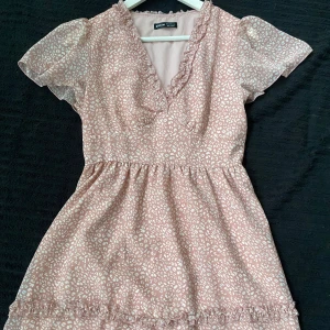 Rosa kortklänning med volang och v-ringning  - Supersöt rosa kortklänning med vitt mönster, v-ringning och volangdetaljer. Klänningen har korta ärmar i transparent material och markerad midja. Perfekt för vår och sommar!