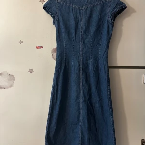 Jeansklänning med knappar från CREEKS . XS/S - Säljer en klassisk jeansklänning från CREEKS i blå denim med korta ärmar och knappar hela vägen framtill. Klänningen har två bröstfickor och en snygg krage. Perfekt för dig som gillar en tidlös och avslappnad stil.