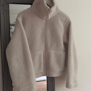 Beige teddyjacka i XS med dragkedja - Supermjuk och fluffig beige teddyjacka i storlek XS. Jackan har hög krage, två stora fickor framtill och stängs med dragkedja. Perfekt för kyliga dagar när du vill vara både varm och snygg. Enkel och stilren design som är lätt att matcha.