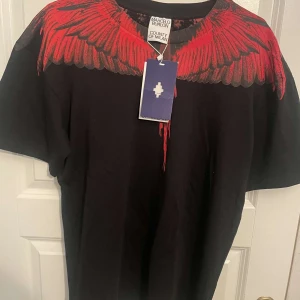 Svart t-shirt Marcelo Burlon med röd print - Svart t-shirt från Marcelo Burlon County of Milan med ikoniskt rött vingmönster över axlar och halsringning. Klassisk passform och korta ärmar. Grym statement-piece för dig som gillar streetwear vibes.