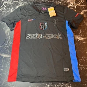 Svart FC Barcelona x Nike fotbollströja - Säljer en svart FC Barcelona fotbollströja från Nike med röda och blå sidopaneler. Tröjan har klubbmärke på bröstet, Nike-logga och tryck med 'Cactus Jack'. UNHCR-logga på ryggen. Tillverkad i Dri-FIT-material för bra ventilation.
