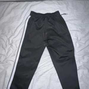 Svarta träningsbyxor från Palm Angels - Svarta träningsbyxor från Palm Angels med vit logga på ena benet och vita ränder längs sidorna. Byxorna har resår i midjan med dragsko och är tillverkade i ett glansigt syntetmaterial. Perfekta för en sportig och trendig look.