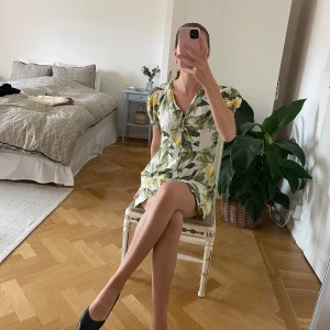 Kort klänning med citronmönster - Säljer en söt kort klänning med citron- och bladmönster i grönt och gult på vit botten. Klänningen har korta ärmar, v-ringning och en avslappnad passform. Perfekt för varma dagar och ger en fräsch sommarkänsla.