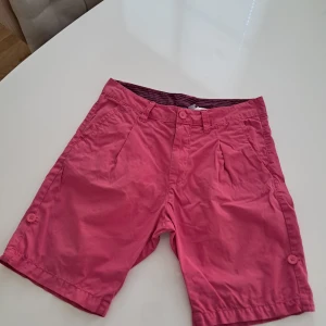 Rosa shorts från Divided, storlek 30 - Snygga rosa shorts från Divided med klassisk passform och knappar i samma färg. Shortsen har fickor fram och bak samt bälteshällor. Tillverkade i bomull, perfekta för varma dagar. Insidan har coola ränder i mörkare rosa. Enkelt och stilrent plagg för sommaren.