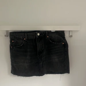 Svart jeanskjol med rå kant - Säljer en svart jeanskjol med klassisk femficksdesign och rå, fransig nederkant. Kjolen har bälteshällor och knappstängning framtill. Perfekt för dig som gillar en enkel men edgy stil.