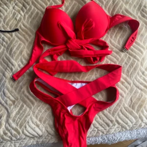 Röd bikini med knyt och snörning - Snygg röd bikini med vadderad topp och breda axelband. Toppen har knyt framtill och underdelen har flera band som korsas över magen för en trendig look. Perfekt för stranden eller poolen. Aldrig använda.