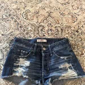 Mörkblå jeansshorts från Hollister - Säljer ett par mörkblå jeansshorts från Hollister med slitna detaljer och fransig kant. Klassisk femficksmodell med låg midja och råa kanter. Perfekta för sommaren!