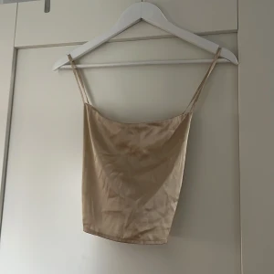Beige croppad linnetopp från NA-KD - Säljer en beige croppad linnetopp från NA-KD med smala axelband och smockad rygg. Toppen har en slät framsida och tunna axelband som korsas i ryggen. 