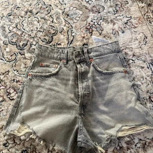 Grå jeansshorts med fransar från Zara - Säljer ett par grå jeansshorts från Zara med råa fransade kanter och klassisk femficksdesign. Shortsen har hög midja och slitna detaljer framtill och baktill för en cool look.