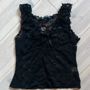 Svart spetslinne med rosett - Säljer ett svart ärmlöst linne i spets med v-ringning och en liten rosett framtill. Toppen har fina spetsdetaljer och är inte använd mycket! Suuuuperfin på fest🥰