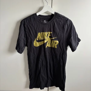 Svart Nike Air t-shirt med guldtryck - Säljer en svart t-shirt från Nike med stort guldigt 'Nike Air'-tryck på bröstet. Klassisk passform och rund halsringning. Perfekt för dig som gillar sportig stil och vill sticka ut med en snygg detalj.