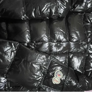moncler maya - Säljer en glansig svart dunjacka från Moncler med klassisk logotyp på ärmen. Jackan har lång ärm, dragkedja och knappar framtill samt en hög krage. Perfekt för kyliga dagar och riktigt snygg streetstil.