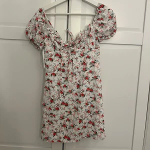 Vit blommig kortklänning med puffärmar - Superfin vit kortklänning med röda och rosa blommor, puffiga kortärmar och söt knytdetalj framtill. Perfekt för sommarens alla tillfällen och ger en romantisk känsla med sitt blommiga mönster och feminina passform.