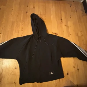 Svart hoodie med dragkedja från Adidas - Svart hoodie från Adidas med dragkedja framtill och klassiska vita ränder längs ärmarna. Tröjan har huva och två fickor framtill. Perfekt för en sportig look eller till vardags.