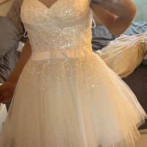 Vit paljettklänning med rosett från Sherri Hill - Superfin vit klänning från Sherri Hill med paljetter och tyllkjol. Klänningen har en markerad midja med bred satinrosett och v-ringad överdel. Perfekt för dig som vill sticka ut med glittriga detaljer och romantisk känsla.