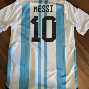 Argentina Messi 10 fotbollströja - Säljer en klassisk Argentina fotbollströja med Messi och nummer 10 på ryggen. Tröjan är vit med ljusblå ränder, svarta detaljer och broderade märken på bröstet. Adidas-logga och AFA-emblem framtill. Perfekt för fotbollsfans!