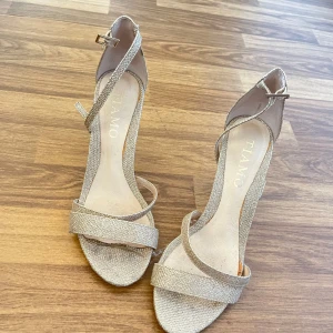 Glittriga klackskor från TIAMO - Säljer ett par glittriga klackar från TIAMO i storlek 37. Skorna har öppen tå, smala remmar och en elegant klack. Perfekta skor som passar till finare tillfällen exempelvis bal. Färg: beige/guld.