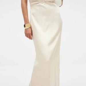 Champagnefärgad långklänning i satin - Elegant slutsåld långklänning i champagnefärgad satin med öppen rygg och slits bak. Klänningen har en smickrande passform och lätt rynkning framtill vid bysten. Perfekt för bal eller fest. Aldrig använd, prislapp finns kvar.