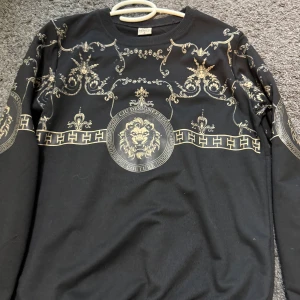 Svart långärmad tröja med lejontryck från Parotti - Svart långärmad tröja från Parotti med stora guldiga lejontryck och dekorativa mönster på bröstet och ärmarna. Tröjan har rund halsringning och detaljerade tryck som ger en lyxig känsla.