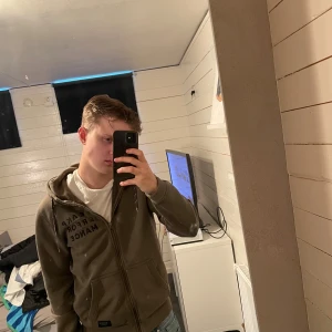 Olivgrön hoodie från Peak Performance - Säljer en olivgrön hoodie från Peak Performance med dragkedja och huva. Tröjan har snörning i huvan och tryckt logga på bröstet. Praktiska fickor framtill och klassisk passform. Perfekt för en avslappnad stil.