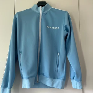 Ljusblå track jacket från Palm Angels - Säljer en ljusblå track jacket från Palm Angels med vit dragkedja och logga på bröstet. Jackan har ståkrage, två fickor med dragkedja och ribbade muddar. Perfekt för en sportig och trendig look. Pris kan diskuteras vid snabb affär 