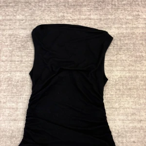 Svart armlös topp - Säljer en svart armlös topp med tight passform. Toppen har rynkade sidor som ger en snygg siluett och är tillverkad i ett mjukt, stretchigt material. Perfekt för dig som vill ha en stilren och trendig look.