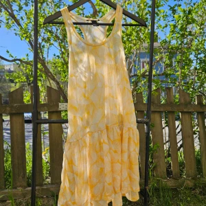 Gulvit sommarklänning med volang - Söt gulvit klänning med bladmönster och volang nertill. Ärmlös modell med rund halsringning och lös passform, perfekt för varma dagar. Lätt och luftig, passar till sommarens alla tillfällen.