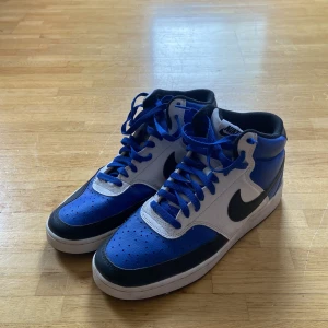 Nike Court Vision Mid blå och vita sneakers - Säljer ett par Nike Court Vision Mid sneakers i blått, vitt och svart. Skorna har klassisk snörning, rund tå och platt sula. Perfekta för dig som gillar streetwear och vill sticka ut med färg. Nike-logga på sidan och bak.