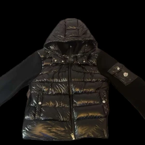 XL men passar på L  - Säljer en svart pufferjacka från Moncler med glansig yta och huva. Jackan har långa ärmar i stickat material, dragkedja framtill och fickor med tryckknappar. Moncler-logga på ärmen och quiltad design på kroppen.