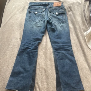  bootcut jeans från True Religion - Säljer ett par blå bootcut jeans från True Religion med slitningar och hål på framsidan. Jeansen har klassiska kontrastsömmar, fickor och knappar framtill. Snygga detaljer på bakfickorna och en cool vintagekänsla.