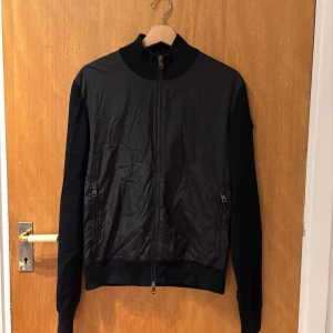 Svart jacka från Moncler med dragkedja - Snygg äkta svart jacka från Moncler med ribbade ärmar och detaljer. Jackan har dragkedja framtill och två fickor med dragkedja. Klassisk Moncler-logga på ärmen och hög krage. Perfekt för en stilren look.