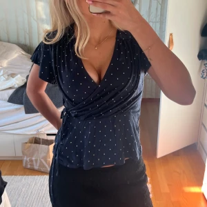 Mörkblå omlottblus med vita prickar - Snygg mörkblå omlottblus med vita prickar och knyt i midjan. Blusen har korta ärmar som är som en liten volang och v-ringning. Perfekt att matcha med både jeans och kjol. Från Lindex Holly & Whyte🌼
