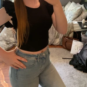 Svart croppad topp med volangärmar - Snygg svart croppad topp med volangdetaljer på axlarna. Rund halsringning och tight passform, perfekt till jeans eller kjol. Enkel men trendig design som passar till många tillfällen.