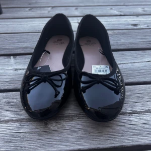 Svarta ballerinaskor med rosett - Säljer ett par glansiga svarta ballerinaskor med söt rosettdetalj framtill. Skorna har rund tå och platt sula, perfekta för en stilren och klassisk look. Passar till många olika outfits och är riktigt bekväma.