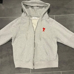 Grå hoodie med dragkedja från Ami - Säljer en grå hoodie från Ami med dragkedja och huva. Jackan har en röd broderad logga på bröstet, två fickor fram och ribbade muddar. Perfekt för en avslappnad stil.