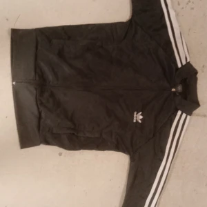 Svart Adidas track jacket med vita ränder - Klassisk svart Adidas track jacket med vita ränder längs ärmarna och broderad logga på bröstet. Jackan har dragkedja framtill och två sidofickor. Perfekt för en sportig och avslappnad stil.