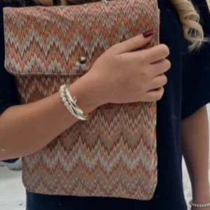 Mönstrad kuvertväska i tyg - Säljer en snygg datorfodral med zigzag-mönster i beige, orange och vitt. Fodralet har ett lock med knappstängning och är perfekt för att bära med sig det viktigaste. Enkel och trendig accessoar som lyfter din outfit.