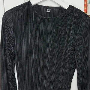 Svart plisserad långklänning - Säljer en svart långklänning med plisserat tyg och rund halsringning. Klänningen har långa ärmar och en elegant, rak siluett som ger ett stilrent intryck. Perfekt för dig som gillar minimalistisk design.