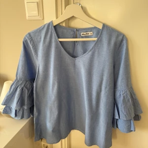 Miss Mille blå blus, storlek M - •Ljusblå blus med V-ringning och trekvartsärmar med volangdetaljer.  •Blusen har en liten knappöppning i nacken. •Använd 2 gånger, som ny. Tvättad. •Djurfritt och rökfritt hem.