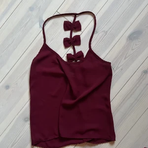Vinröd linnetopp med rosetter från SheIn - Supersöt vinröd linnetopp från SheIn med smala axelband och tre dekorativa rosetter i ryggen. Perfekt för dig som vill ha en enkel men ändå unik topp. Tunt och lätt material som passar super till fest. Säljer för att den inte används!🥰