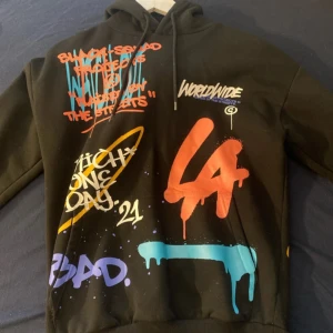 Svart hoodie med färgglada tryck och graffiti-text - Cool svart hoodie med färgglada graffiti-inspirerade tryck och texter som 'LA', 'Worldwide', 'Drip2Hard' och 'Rich One Day'. Hoodien har huva med dragsko och tryck på både fram- och baksida samt ärmar. Perfekt för dig som gillar streetwear och unika plagg.