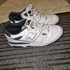 Vita New Balance 550 sneakers - Säljer ett par klassiska vita New Balance 550 sneakers med svarta detaljer. Skorna har snörning och en platt sula, perfekt för en sportig och stilren look. Ikonisk N-logga på sidan och 550-tryck. Passar till många outfits!