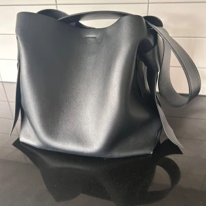  Acne Studios Musubi Midi Bag - Musubi Midi Bag från Acne Studios med dammpåse.  Detta är en av märkets signaturmodeller, tillverkad av 100 % premiumläder. Levereras med en justerbar axelrem för mångsidig användning.  Inuti har väskan en ficka med dragkedja och två öppna fack för enkel organisering.  Den diskreta logotypen är präglad på framsidan. Toppen stängs säkert med en magnetisk stängning.              