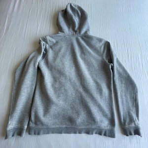 Ljusgrå hoodie från Sail Racing - Ljusgrå hoodie med dragkedja från Sail Racing. Tröjan har huva, långa ärmar och två fickor framtill. Perfekt för en avslappnad stil och enkel att matcha med det mesta.