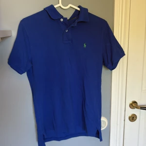 Blå pikétröja från Polo Ralph Lauren - Säljer en klassisk blå pikétröja från Polo Ralph Lauren med krage och knappslå. Tröjan har korta ärmar och den ikoniska gröna loggan broderad på bröstet. Perfekt för en stilren och avslappnad look. Storleken är 14-16 år Large