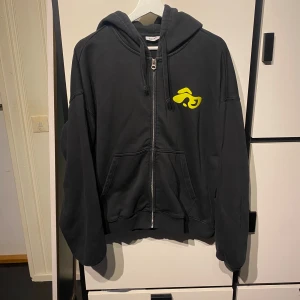 Svart hoodie med gul graffiti från Weekday - Säljer en svart hoodie från Weekday med dragkedja och huva. Framtill finns en gul graffiti-liknande logga på bröstet och en större på ryggen. Tröjan har fickor framtill och en avslappnad passform.