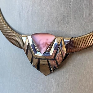 Vintage halsband med rosa detalj - Unikt vintage halsband i silverfärgad metall med en geometrisk design och en stor, triangelformad lila detalj i mitten. Halsbandet har en bred, ormliknande kedja och stängs med ett klassiskt spänne. Perfekt för dig som vill sticka ut med ett statement-smycke.