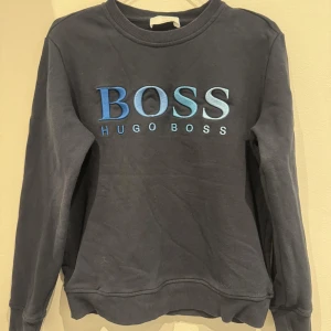 Svart sweatshirt från Hugo Boss - Säljer en svart sweatshirt från Hugo Boss med stor broderad logga i blå och turkosa nyanser på bröstet. Tröjan har rund halsringning, långa ärmar och ribbade muddar vid ärmslut och nederkant. Perfekt för en stilren och avslappnad look.
