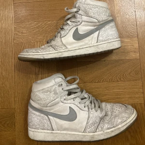 Nike Air Jordan 1’s med grå detaljer och mönster - Säljer ett par vita Nike Air Jordans med grå swoosh och unikt mönster över hela skon. Högt skaft, snörning och rund tå. Perfekta för dig som vill sticka ut med stilrena sneakers.