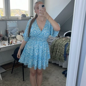 Blåvit omlottklänning med mönster - Säljer en blåvit kortklänning med omlottmodell och v-ringning. Klänningen har korta, vida ärmar och ett fint blommigt mönster. Resår i midjan ger en smickrande passform och kjolen har lätt volang. Perfekt för sommarens alla tillfällen!